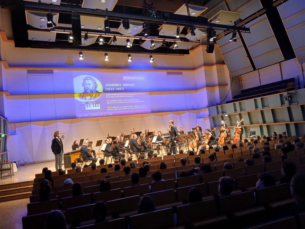 Kuva Taidetestaajat-konsertista. Yleisöä ja orkesteri. Taustalla näkyy dia jossa kerrotaan säveltäjästä.